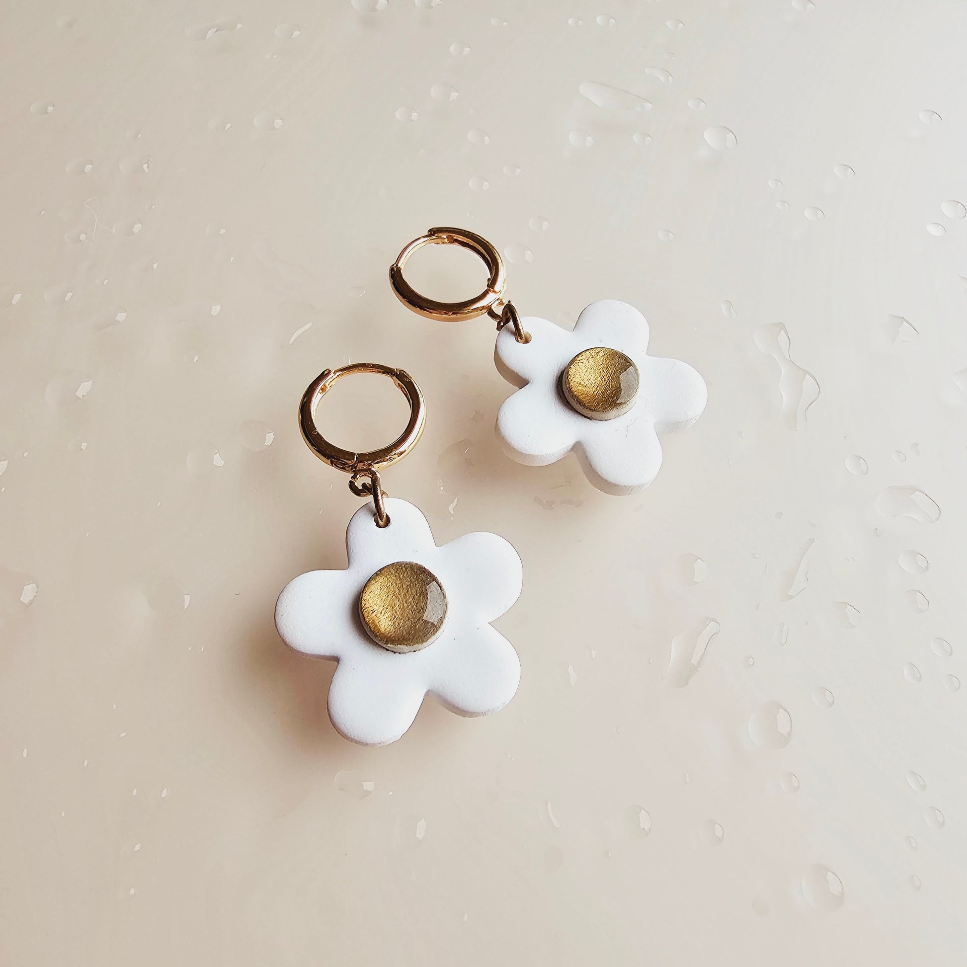 The image shows 2 daisy white earrings. The earrings are a white flower with a gold centre hanging from a gold push back hoop. The daisy flowers are made from polymer clay.  Evelet Designs. 