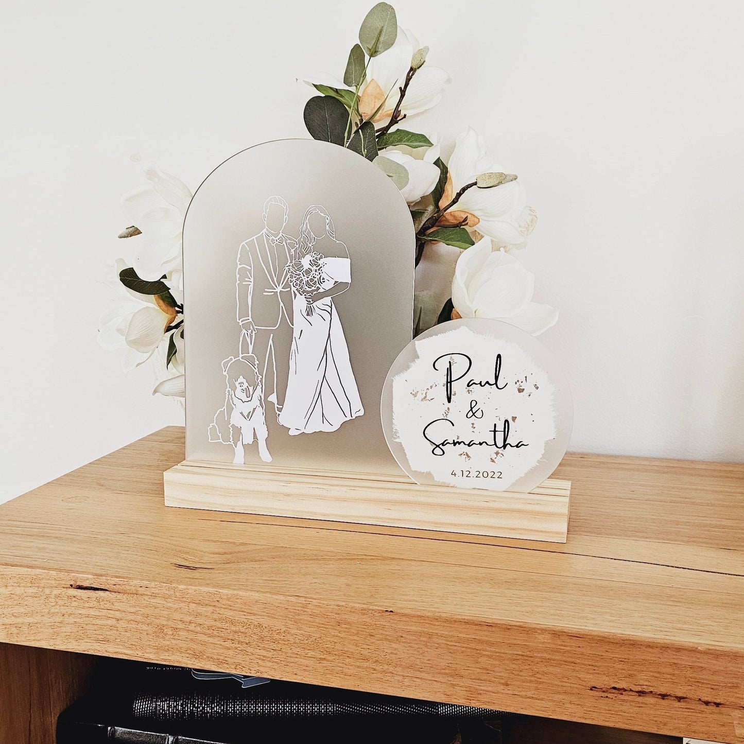 This image shows an illustration on an arch. The illustration is in white vinyl and shows a dog, a groom and a bride in a white wedding dress. The arch is sitting in a pine timber base. There is a circle acrylic shape next to it that says "Paul and Samantha" and the date 4.12.2022. It is perfect as a wedding or anniversary gift