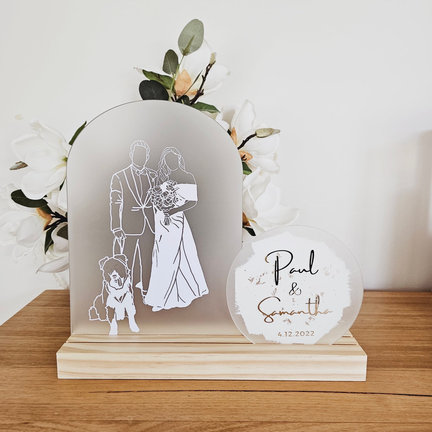 This image shows an illustration on an arch. The illustration is in white vinyl and shows a dog, a groom and a bride in a white wedding dress. The arch is sitting in a pine timber base. There is a circle acrylic shape next to it that says "Paul and Samantha" and the date 4.12.2022.
